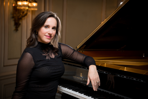 A woman with brown hair in a black sheer performance shirt sits with her elbow on a grand piano