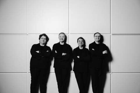Black-and-white photo of four men dressed in all black smiling in front of a geometric square light background