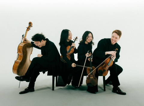 A string quartet in all-black sit in different directions on a bench in front of a white-blue ombré background