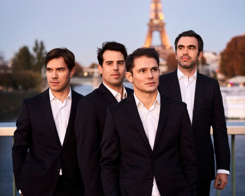 Four men in matching black suits with white shirts pose in front of the Eiffel tower at sunset