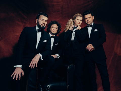 Three men and one woman, all wearing tuxes with bowties, pose for a photo in front of a red background