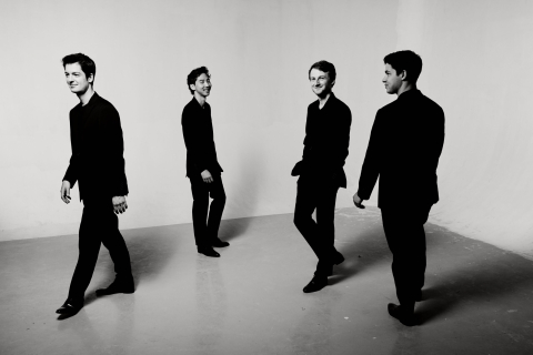 Black-and-white photo of four men in all black spaced out in a room walking