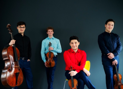 Four men in brightly colored clothes smile and pose with their string instruments in front of a blue background