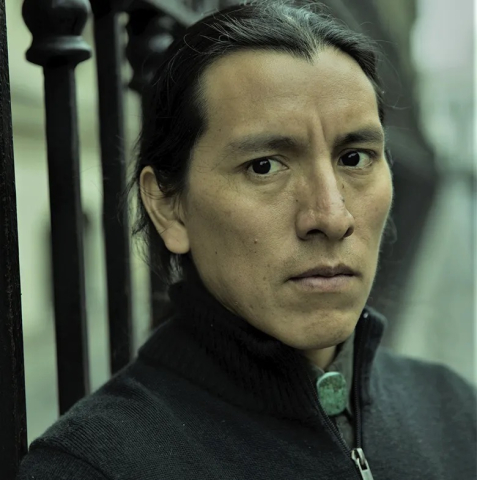 A man looks sternly at the camera while leaning on a black metal fence and wearing a quarter-zip