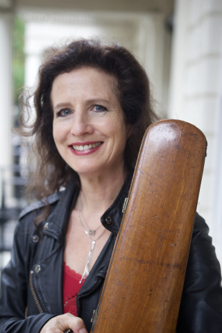 A woman with dark brown hair, a red shirt, and black jacket holds her violin case and smiles at the camera
