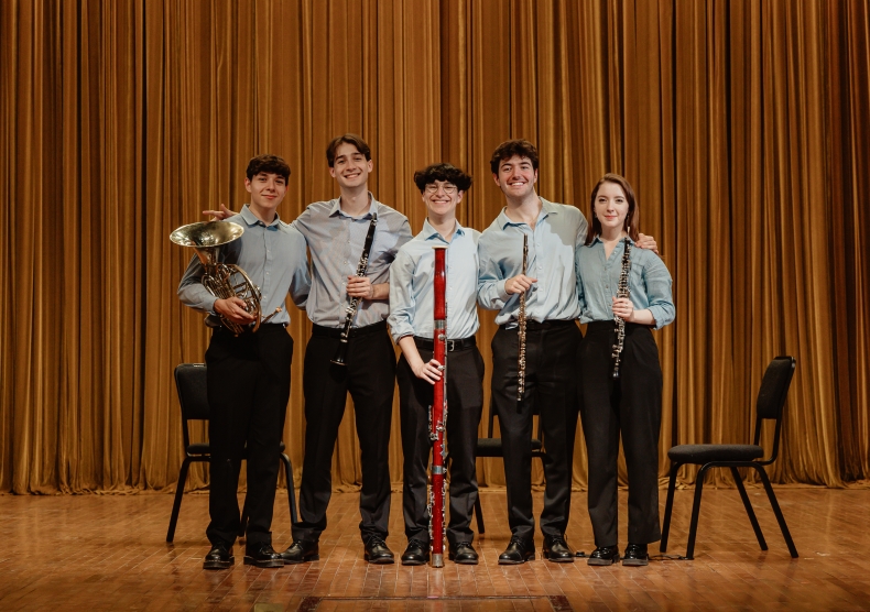 Five students in blue dress shirts and black pants stand side-by-side with their wind instruments and smile for a photo
