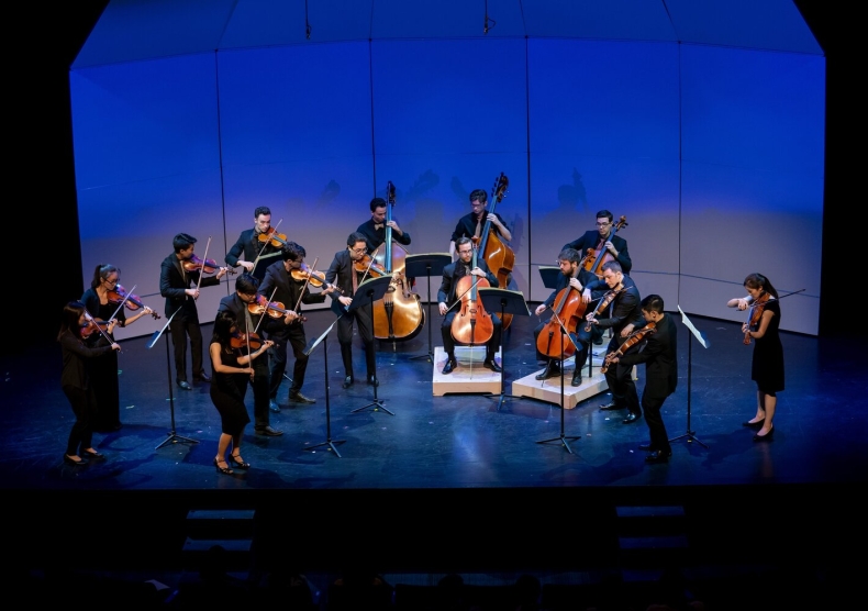 Conductorless string orchestra performs on stage in front of blue lighting