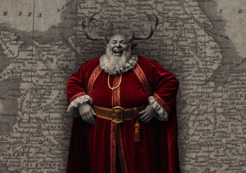 A larger older man in a red robe, with antlers on his head, stands with his hands on his hips in front of a world map