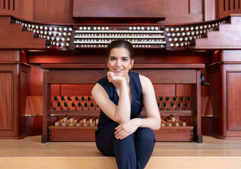 Emily sits on the step in front of the grand organ, smiling with one hand propped under her chin