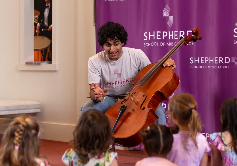 A cellist sitting down in a Shepherd School t-shirt talks about his instrument in front of a group of young kids, whose backs are facing the camera
