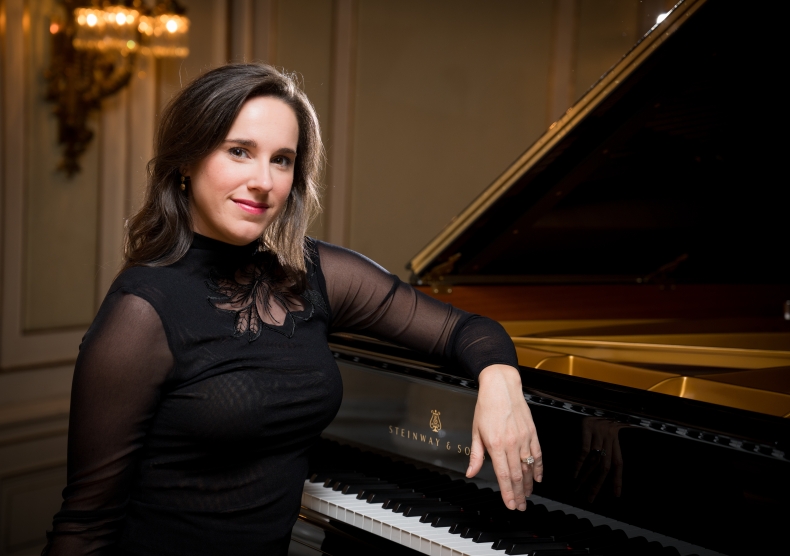 A woman with brown hair in a black sheer performance shirt sits with her elbow on a grand piano