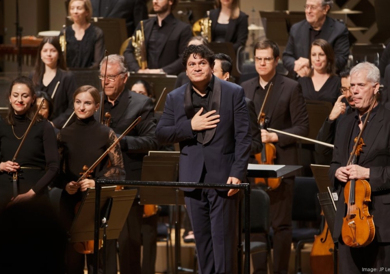 Cristian Măcelaru takes a bow with the Cincinnati Symphony Orchestra in his debut concert. He wears a blue suit with his hand on his chest and a small smile on his face, looking upwards. He stands in front of orchestra musicians dressed in black concert attire.