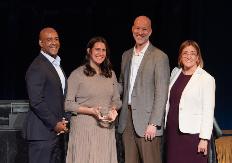 Four people pose for a photo. From left to right, a man in a blue suit, a woman in a beige dress holding an award made out of glass, a man in a tan suit, and a woman in a burgundy dress and white blazer