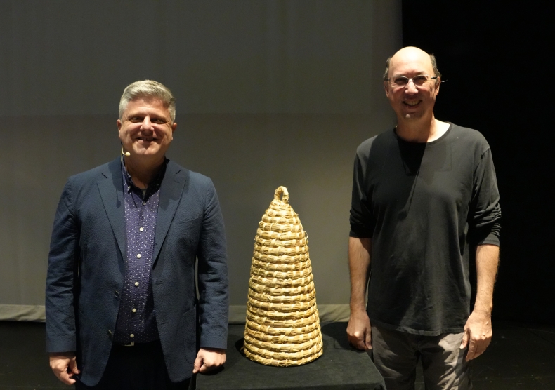 Two smiling men pose with a honeycomb placed on a table between them
