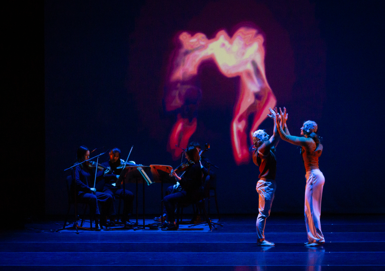 A string trio performs in front of a projected light of fire/smoke as two dancers wearing EEG brain caps press their hands together