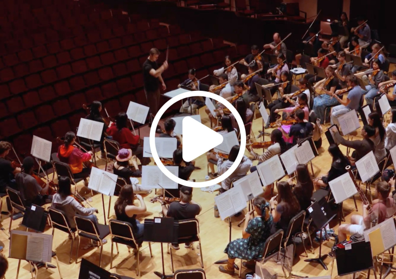 An overhead shot of students in orchestra rehearsal. Image has a play button on it.