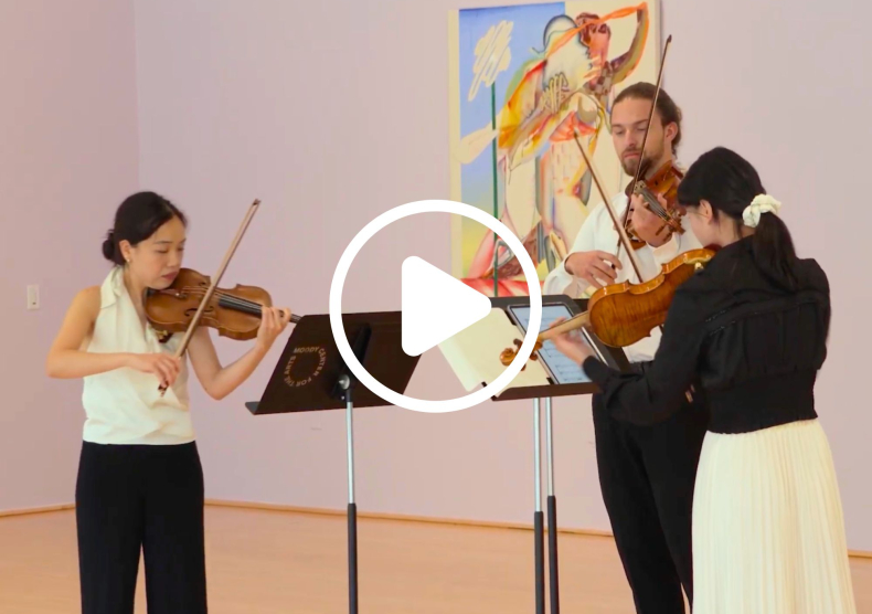 Three string players perform at the Moody Center for the Arts, and are pictured in front of a large painting. The image has a play button on it.