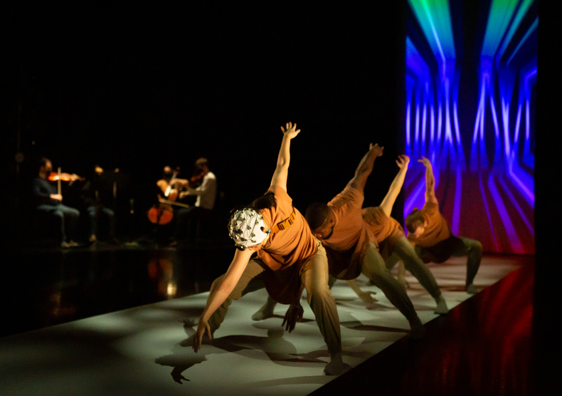 Four dancers pose in a line, with the first wearing a brain cap. In the distance, you can see the shadows of a string quartet performing.