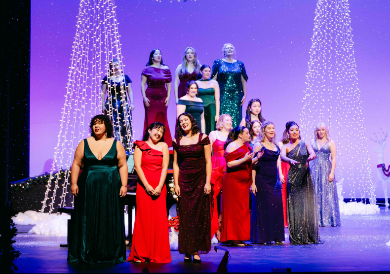 An ensemble of vocalists in long gowns perform on stage in front of a purple background with two Christmas lights made out of twinkle lights