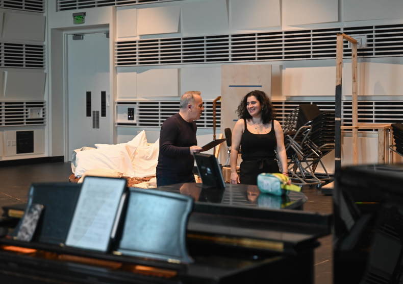 Photo of the stage director working with opera student behind a piano and in front of a bed (set piece)