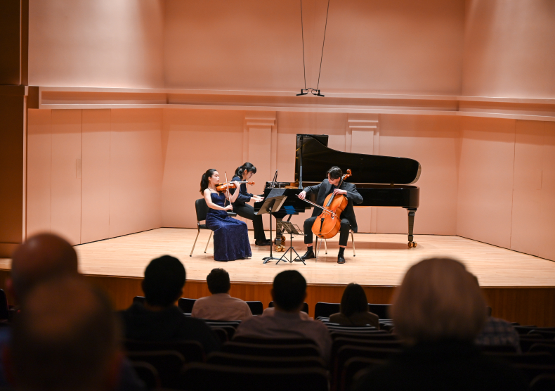 A piano trio performs on the Duncan stage, with the backs of audience heads visible as they watch