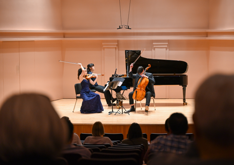 A piano trio performs on the Duncan stage, with the backs of audience heads visible as they watch