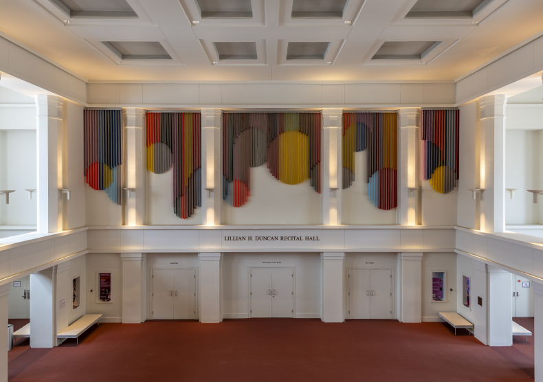 Grand Foyer of Alice Pratt Brown Hall, featuring white walls with sconces, a large-scale art piece with draped arches, and a red carpet