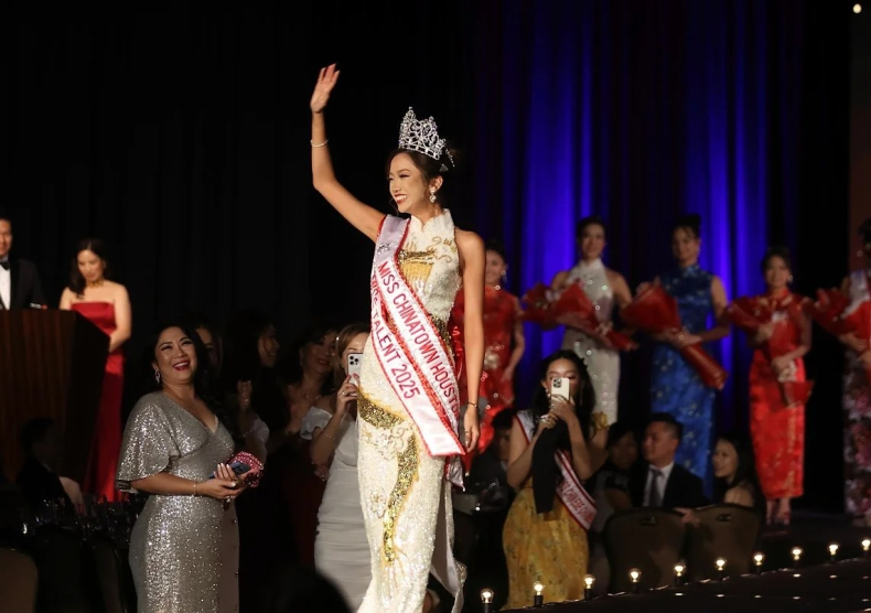 A Chinese-American woman at a pageant waves, she wears a glittery white and gold dress, a silver tiara, and two sashes