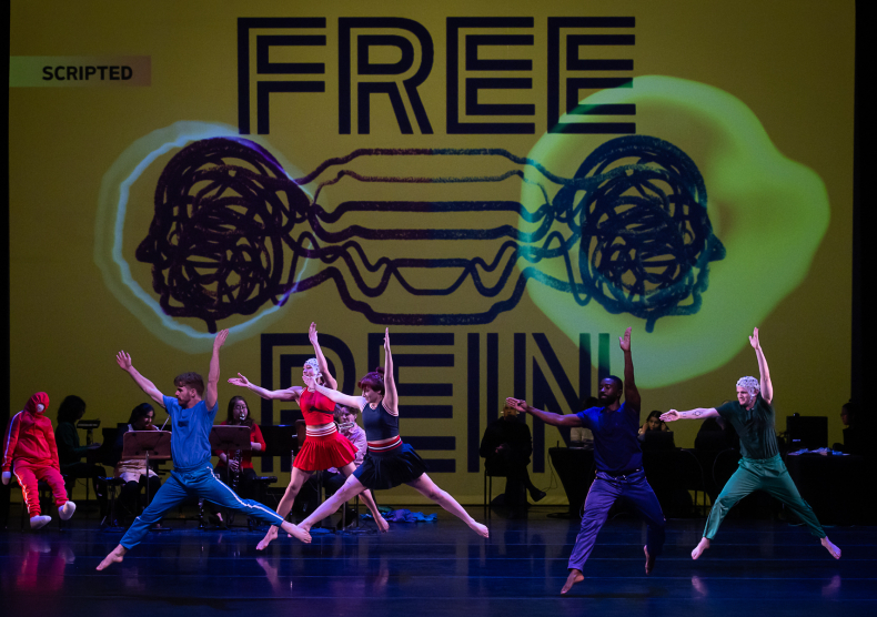 A group of dancers make the same jumping move in the area in front of a screen with graphics and the words "Free Rein"