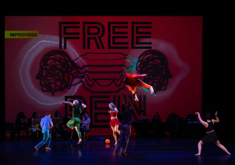 A mannequin flies through the air alongside a group of dancers in front of a red screen with graphics and the words "Free Rein"