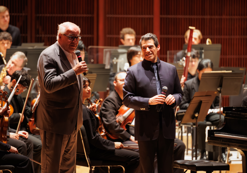 Two older men stand on stage in front of an orchestra; on the left, the man with white hair and a gray jacket speaks with a microphone, while the man on the right smiles at him, wearing a blue suit