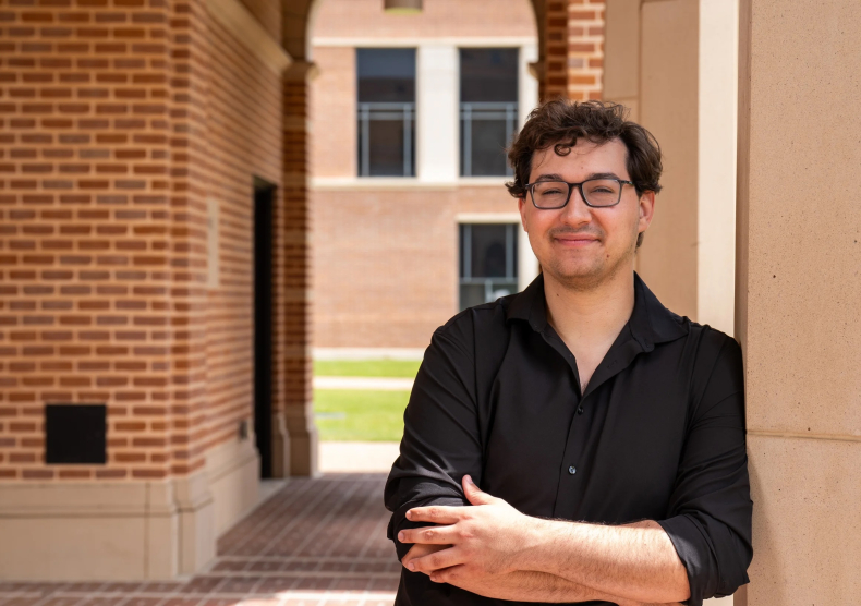 Headshot of a young man wearing a black shirt, glasses, and crossing his arm; he stands in a brick archway