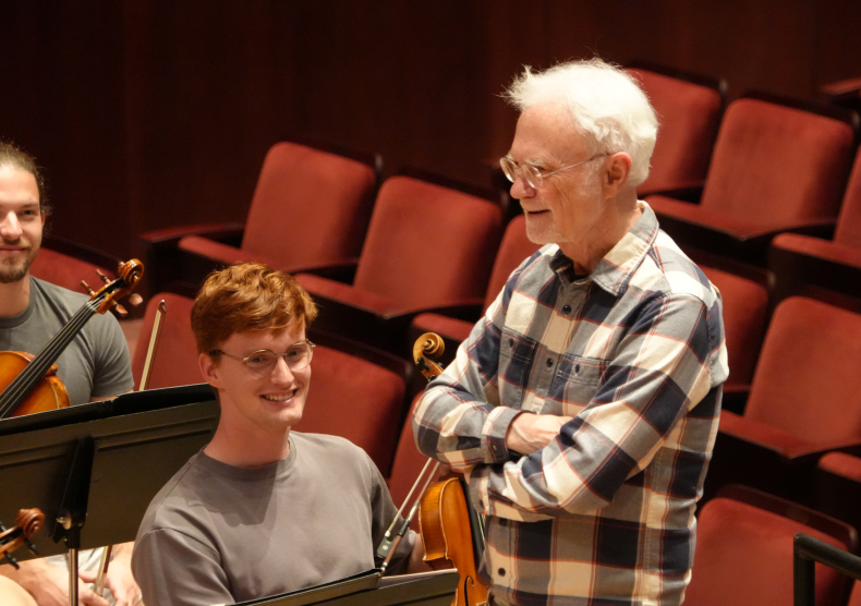 Two young male violists smile in orchestra rehearsal as John Adams, an older man with white hair wearing a plaid shirt, crosses his arms and smiles while standing on the conductor podium