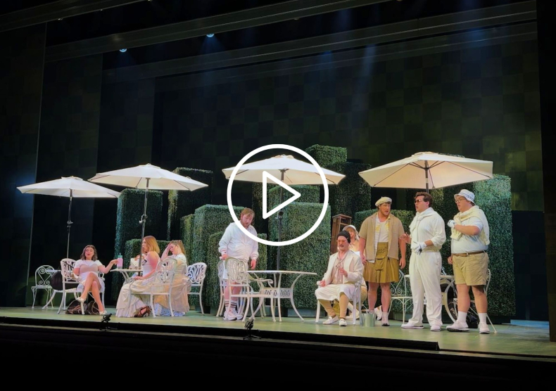 Scene of opera students dressed in white and beige on stage in front of a green set; play button on photo