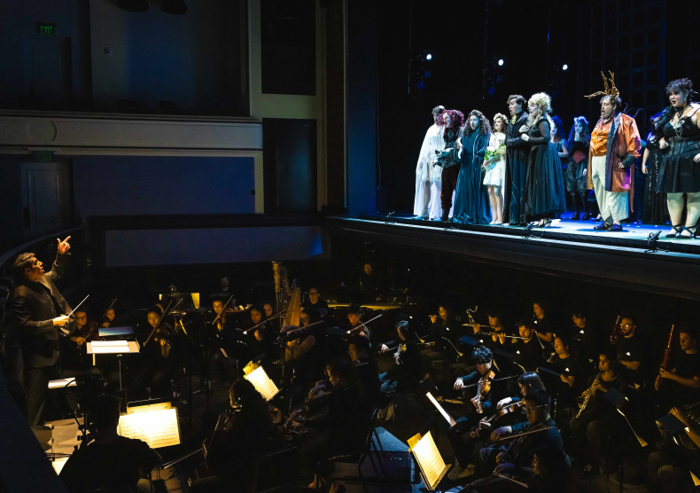 A photo of a conductor leading vocalists on stage in costume and a full orchestra in black clothes in the pit