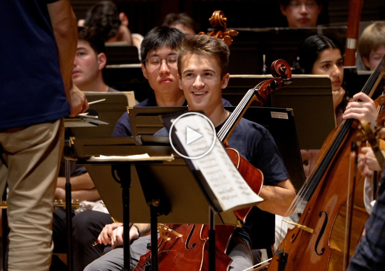 Thumbnail of a cellist smiling in orchestra rehearsal as he looks at the conductor