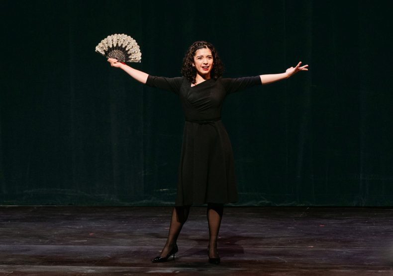 A woman with her arms outstretched and holding a fan sings in front of a black curtain