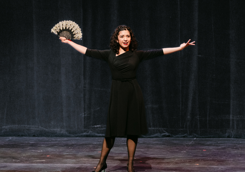 A woman with her arms outstretched and holding a fan sings in front of a black curtain