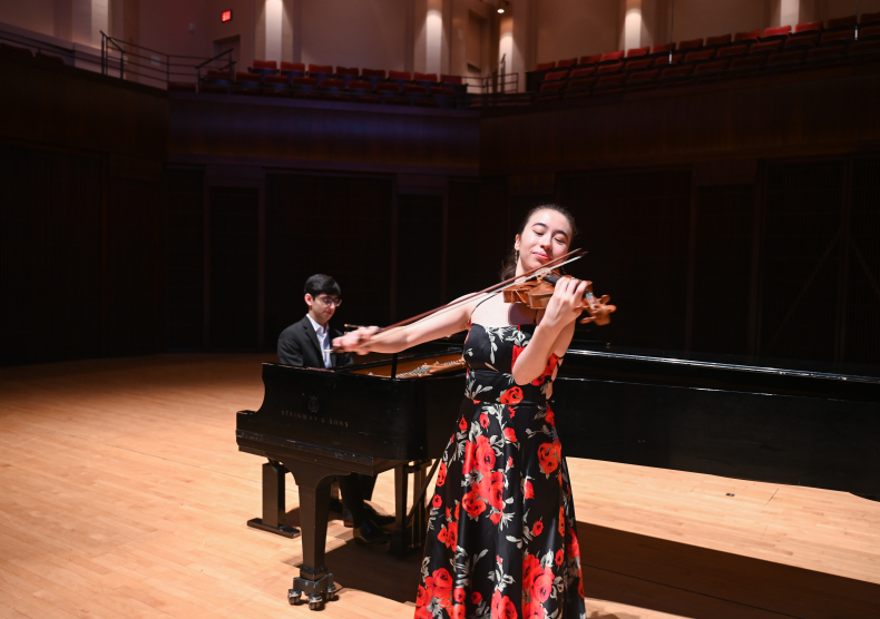 A violinist in a red and black dress performs with a pianist in a suit in Stude Concert Hall