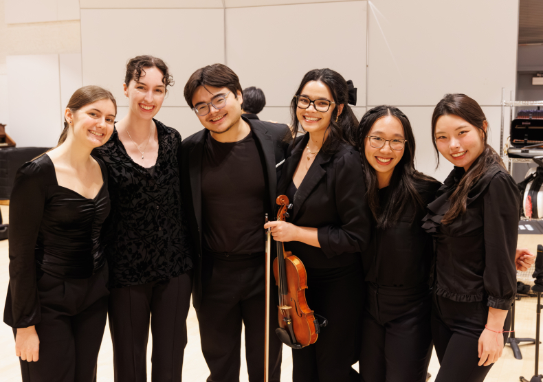 Six students wearing concert black, one holding a violin, smile and pose for a photo backstage