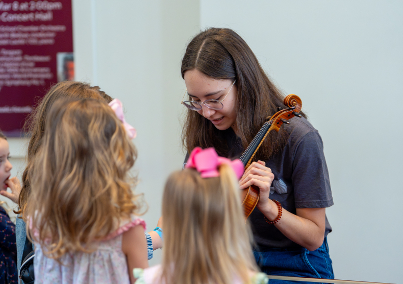 A woman in a grey t-shirt shows off her violin to a few young girls with blonde hair