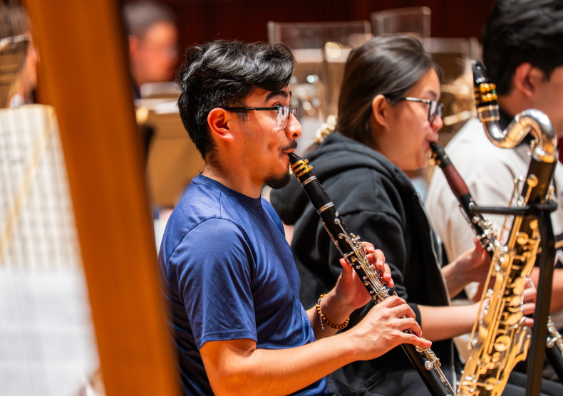 A young man in a blue shirt plays the clarinet in orchestra rehearsal; near him is another young woman playing the clarinet