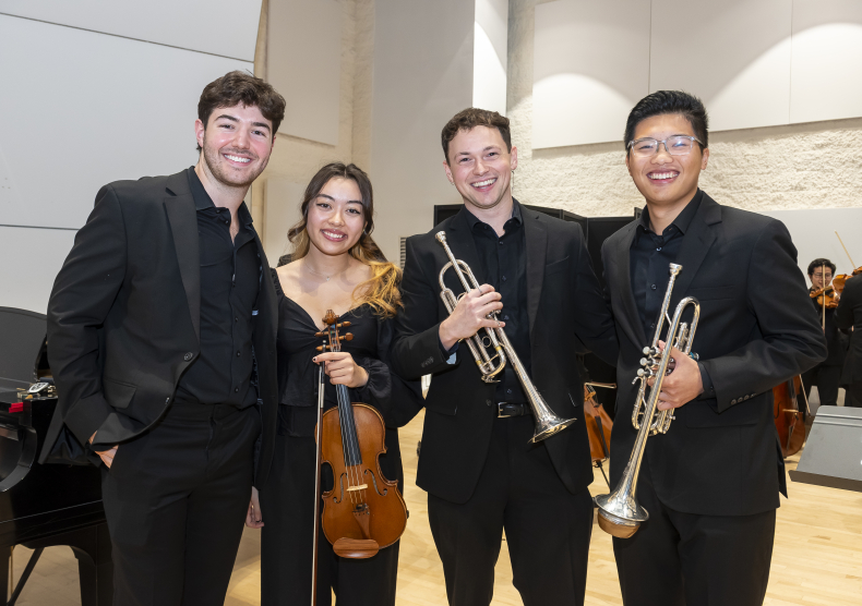 Four students wearing concert black and holding instruments (two trumpets and one violin) smile for a photo