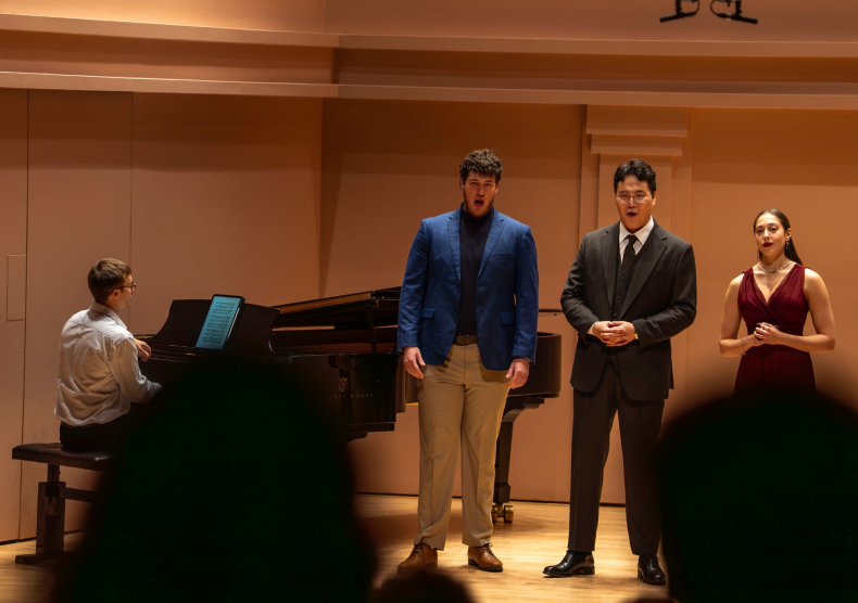 A seated pianist and three standing singers perform on stage in a recital hall