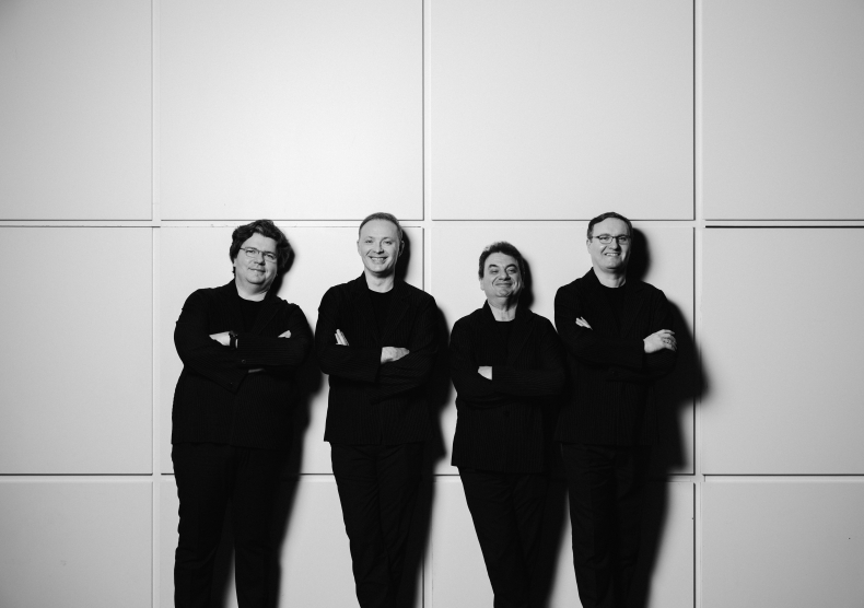 Black-and-white photo of four men dressed in all black smiling in front of a geometric square light background