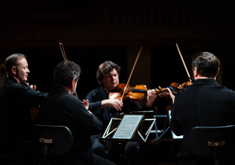 The side view of four men performing in a string quartet, wearing black clothes in front of a black background