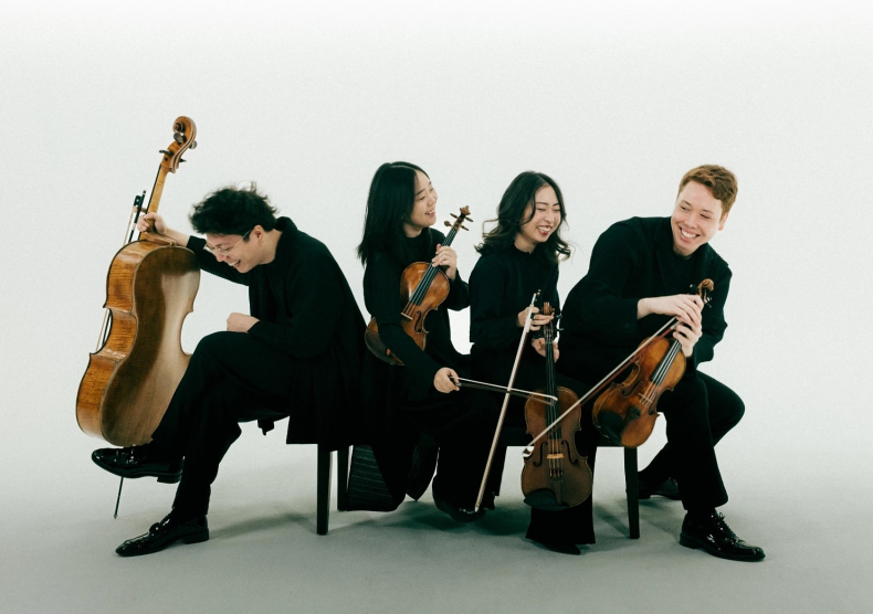 A string quartet in all-black sit in different directions on a bench in front of a white-blue ombré background