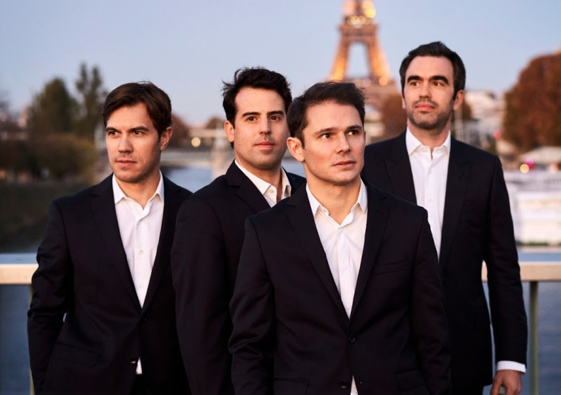 Four men in matching black suits with white shirts pose in front of the Eiffel tower at sunset