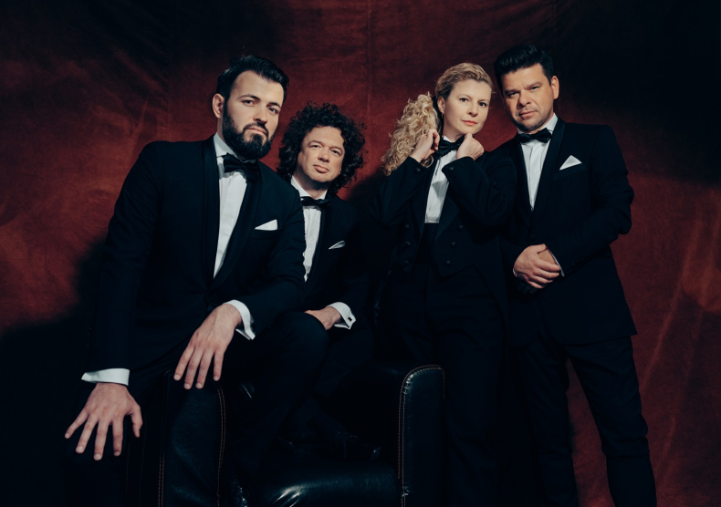 Three men and one woman, all wearing tuxes with bowties, pose for a photo in front of a red background