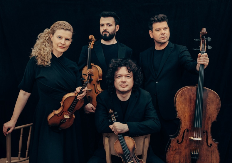 A string quartet in all black poses for a photo, one man sits while the others stand behind him